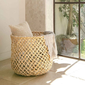 The Kearny Bleached Round Basket by Pigeon & Poodle, filled with a beige textured pillow and light blanket, sits on a stone floor near a large window overlooking a potted tree.