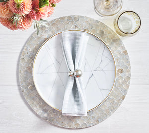 A white plate with a marble pattern sits on the Camellia Gray Placemat by Kim Seybert, topped with a folded gray napkin in a silver ring. Two glasses and a floral arrangement are placed nearby.