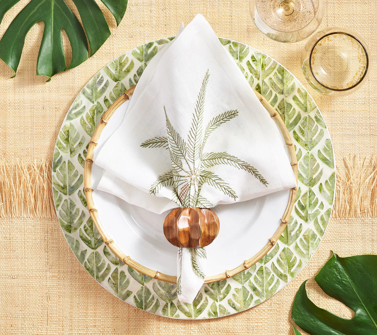 A table setting featuring a tropical leaf-patterned plate, bamboo-style charger, palm tree napkin, Kim Seybert Twist Brown/Gold Napkin Ring, wine glass, green leaf accents on a woven placemat, and gold inlaid brass details for elegance.