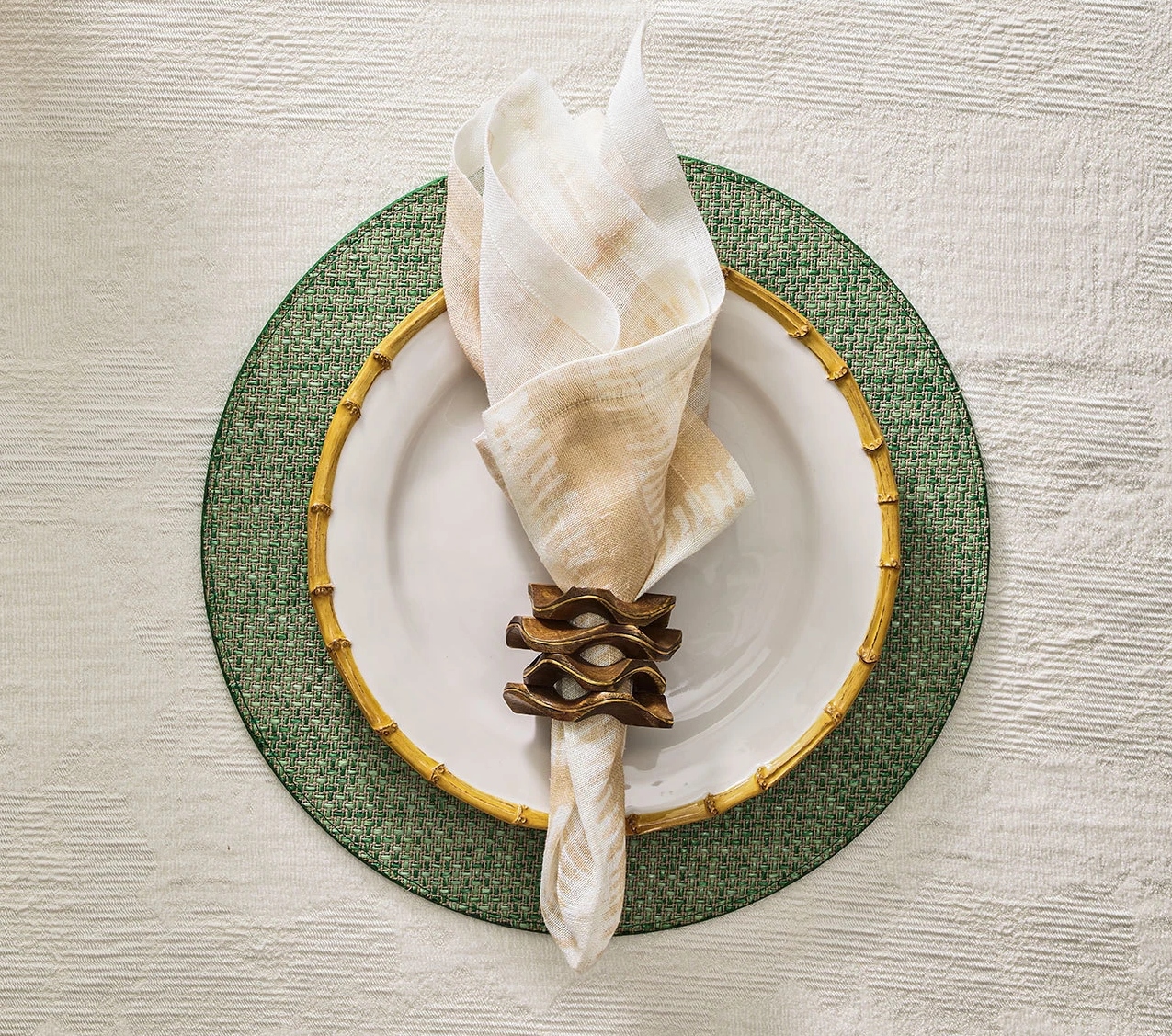 A white plate with a gold bamboo-patterned rim sits on a green charger, topped with a cream napkin held by the Kim Seybert Pavilion Brown/Gold Napkin Ring, featuring hand-carved mango wood and an inlaid brass edge.