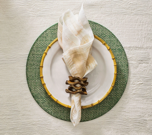A white plate with a gold bamboo-patterned rim sits on a green charger, topped with a cream napkin held by the Kim Seybert Pavilion Brown/Gold Napkin Ring, featuring hand-carved mango wood and an inlaid brass edge.