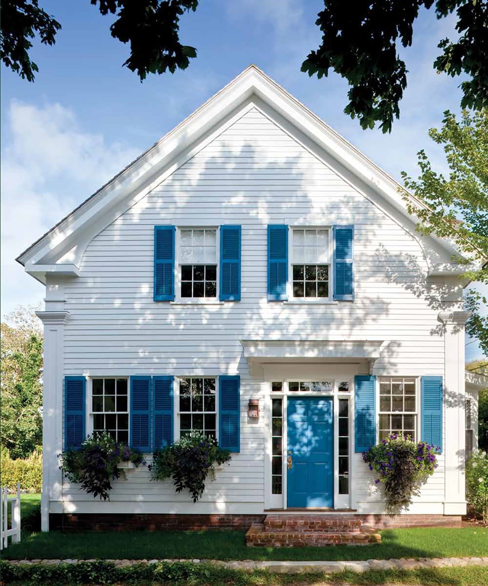 A white two-story classical home with blue shutters and a blue door, framed by trees, features two potted plants beneath the windows. This reflects the site-sensitive philosophy highlighted in "Timeless: Classic American Architecture for Contemporary Living" by Common Ground.
