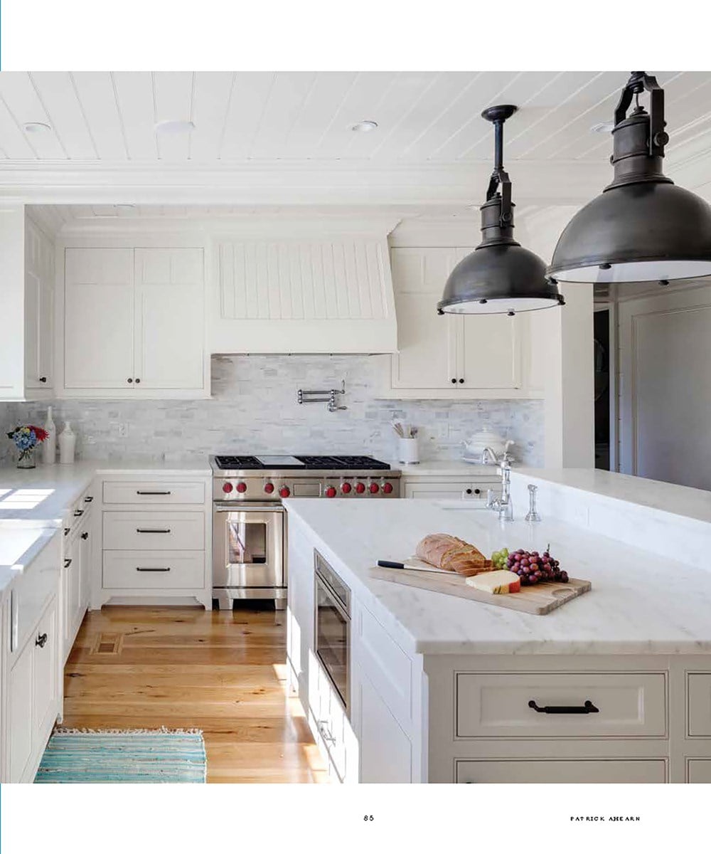 A modern kitchen with white cabinetry, stainless steel appliances, and wooden flooring reflects the essence of Common Ground's "Timeless: Classic American Architecture for Contemporary Living." A loaf of bread and grapes sit on a cutting board beneath two large pendant lights.
