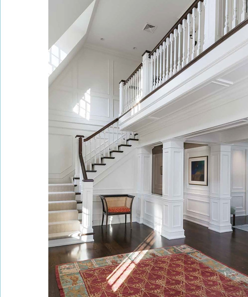 A spacious foyer with a staircase and dark wood floors is enhanced by a colorful rug, white walls, and natural light showcasing a small bench and artwork. Skilled residential designers’ touch echoes the philosophy in "Timeless: Classic American Architecture for Contemporary Living" by Common Ground.