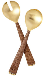 The Ananta Serving Set by Be Home is a handcrafted 2-piece set from India, featuring gold-toned serving utensils with woven brown handles—one slotted spoon and one solid spoon—displayed on a white background.