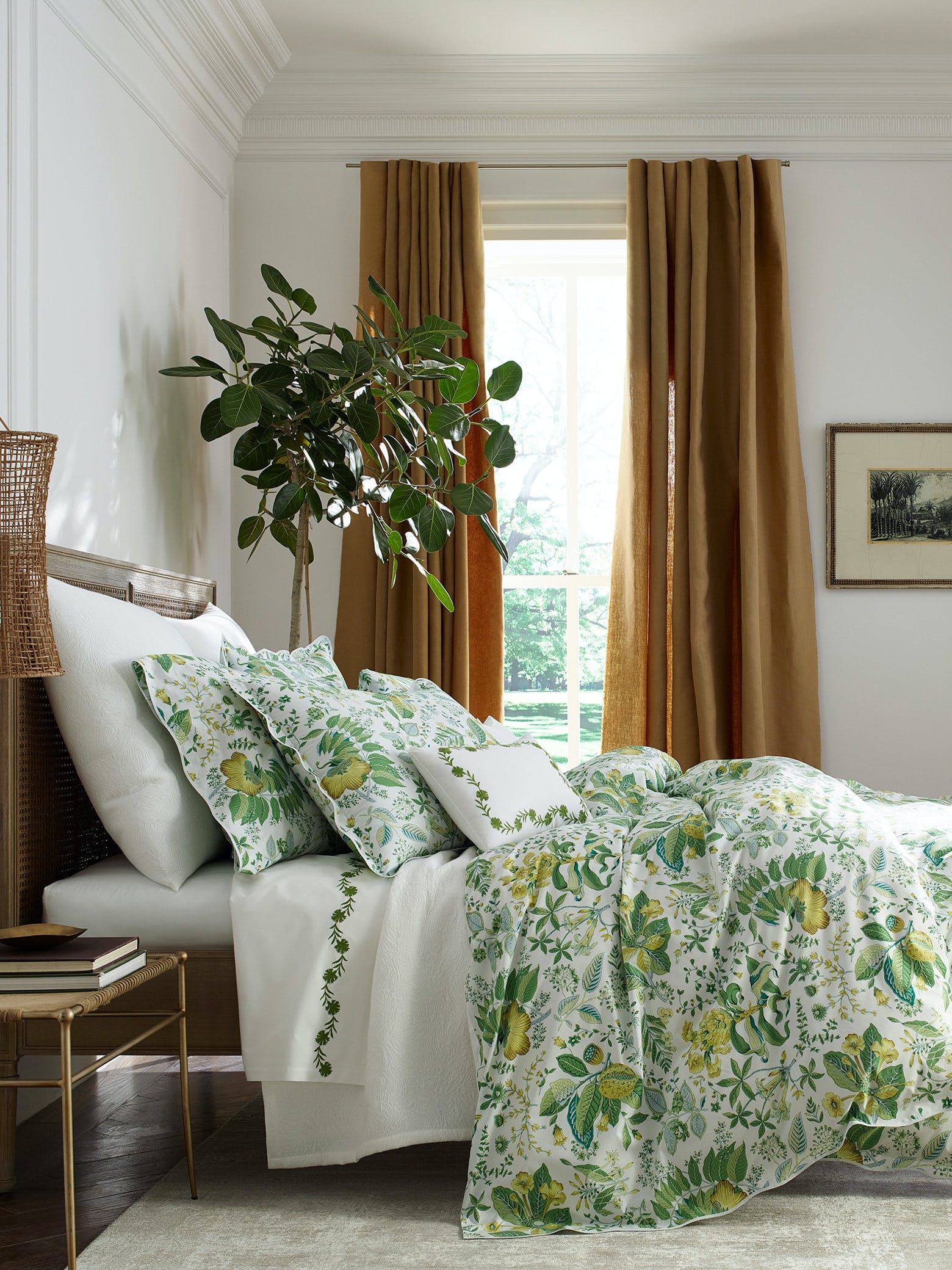 A neatly made bed with Matouk’s Pomegranate Bedding Collection in Citrus, several pillows, a potted plant by the bed, and golden-brown curtains covering tall windows in a bright bedroom.