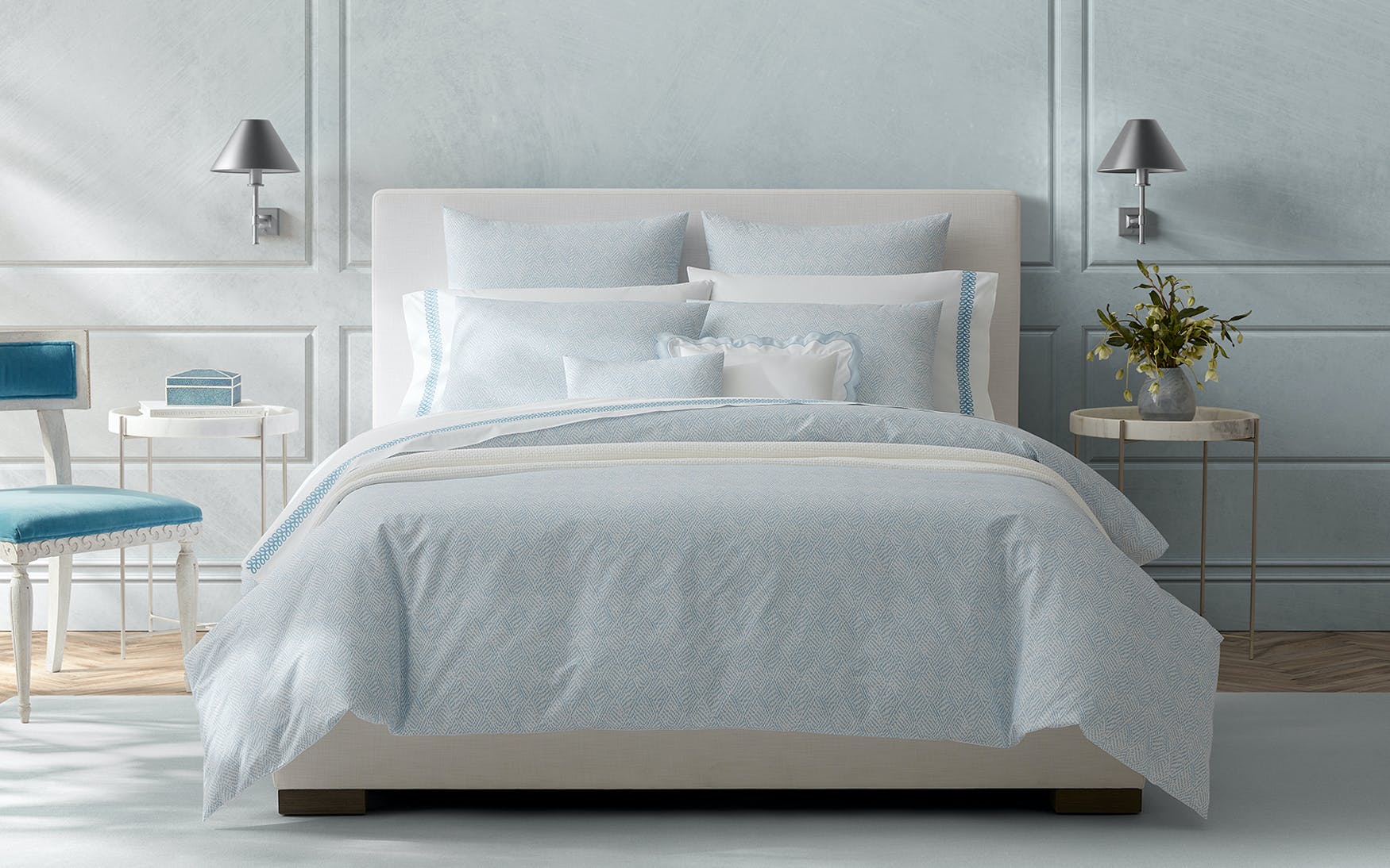 A modern bedroom with the Matouk Duma Diamond Bedding Collection in Sky, featuring light blue and white Egyptian cotton, two round side tables with lamps, a flower vase, and soft geometric accents.
