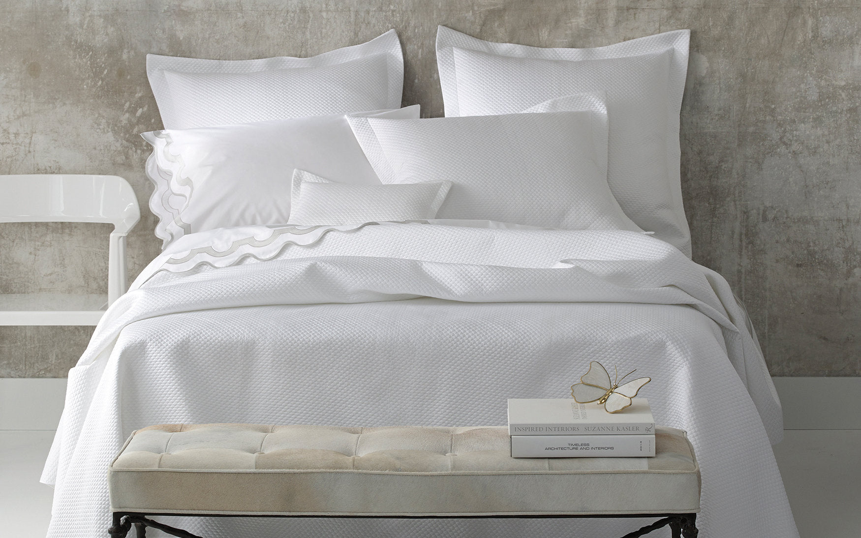 A neatly made bed features the Matouk Alba Bedding Collection in White, with pillows and a small bench at the foot holding books and glasses, all set against a concrete-style wall.