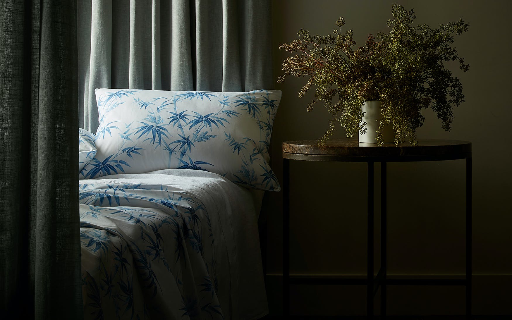 A bed dressed in Matouk's Dominique Bedding Collection, Azure—white and blue Egyptian cotton with bold botanicals—stands by a round wooden table with dried flowers, beside green curtains in soft light.