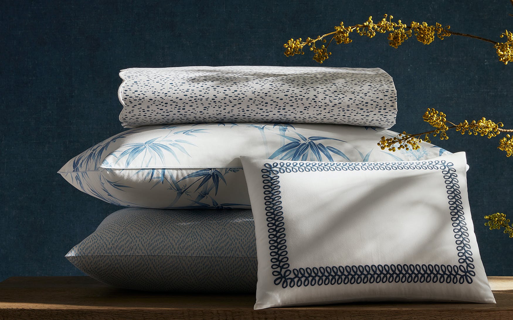 The Matouk Duma Diamond Bedding Collection in Navy, crafted from luxurious 500 thread count Egyptian cotton percale, features folded bedding and pillows with blue and white harlequin patterns on wood, with yellow flowers in the foreground.