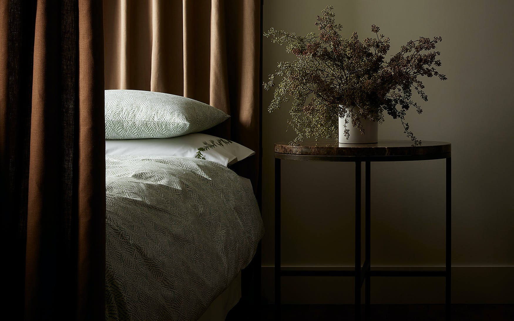 A bedside table with a vase of dried foliage stands next to a bed dressed in Matouk’s Duma Diamond Bedding Collection in Grass, made from OEKO-TEX® certified Egyptian cotton percale, with brown curtains in a dimly lit room.