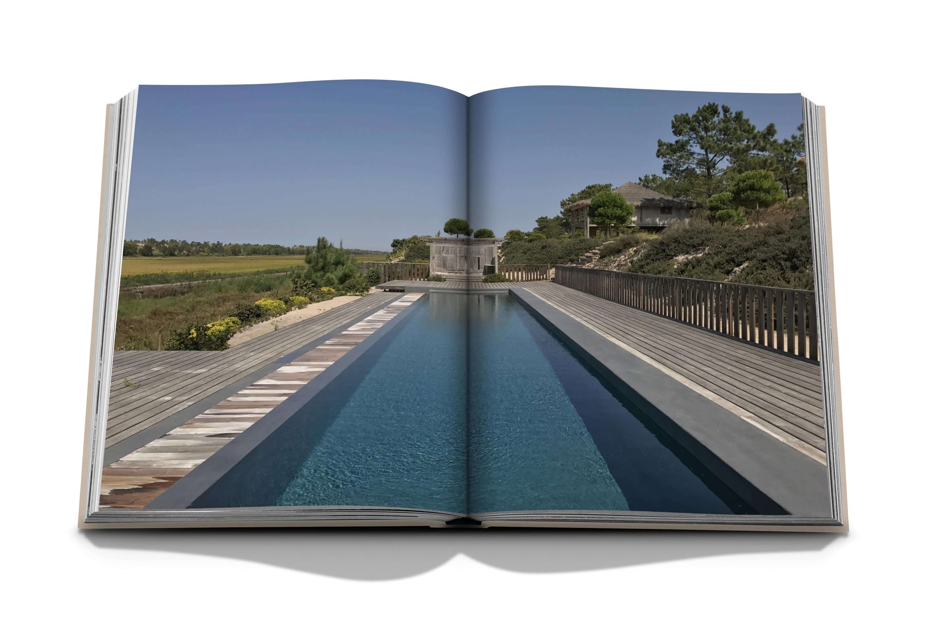 Open book displaying a photograph of a peaceful, rectangular swimming pool with wooden decking, surrounded by greenery and distant buildings under a clear blue sky—reminiscent of the serene beaches of Comporta Bliss, celebrated by its artistic community—by Assouline.