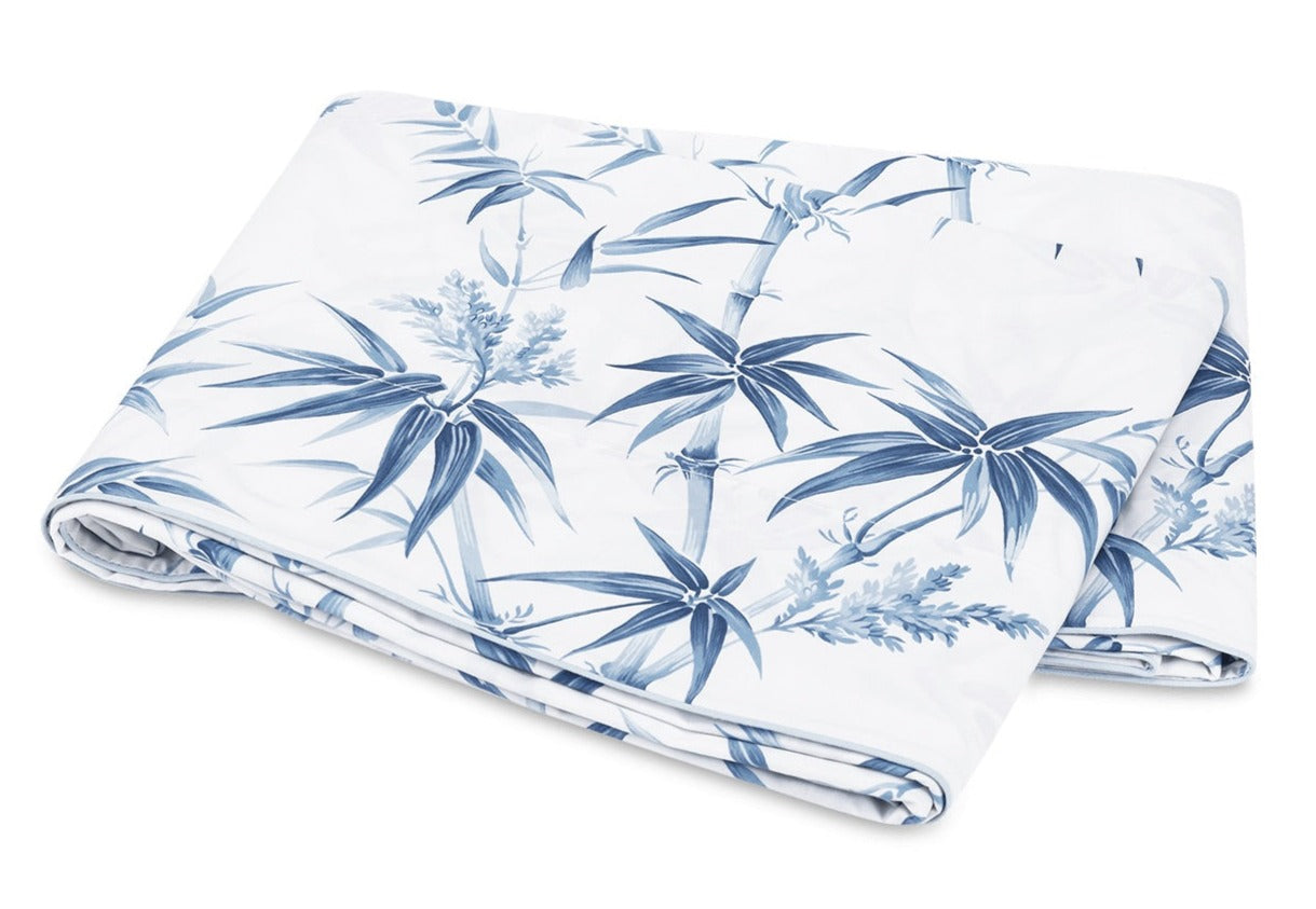A folded white blanket from the Dominique Bedding Collection, Azure by Matouk, featuring a blue tropical leaf and plant pattern and crafted from soft Egyptian cotton.