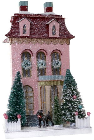 The Pink Chateau by Cody Foster is a pink, snow-covered two-story house with window wreaths, two decorated evergreens, a white fence, and a black dog in front.