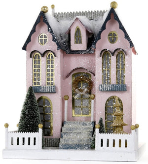 The Pastel Pink Manor by Cody Foster is a 9"W x 10"L Victorian-style paper mache house, featuring gold-trimmed windows, a snowy roof, two small trees, and a white picket fence.