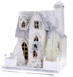 The Cody Foster Frosted Mansionette is a glittery mini paper mache model of a white house with snowy roof, trees, and picket fence, creating a charming winter holiday scene. See product description for dimensions.