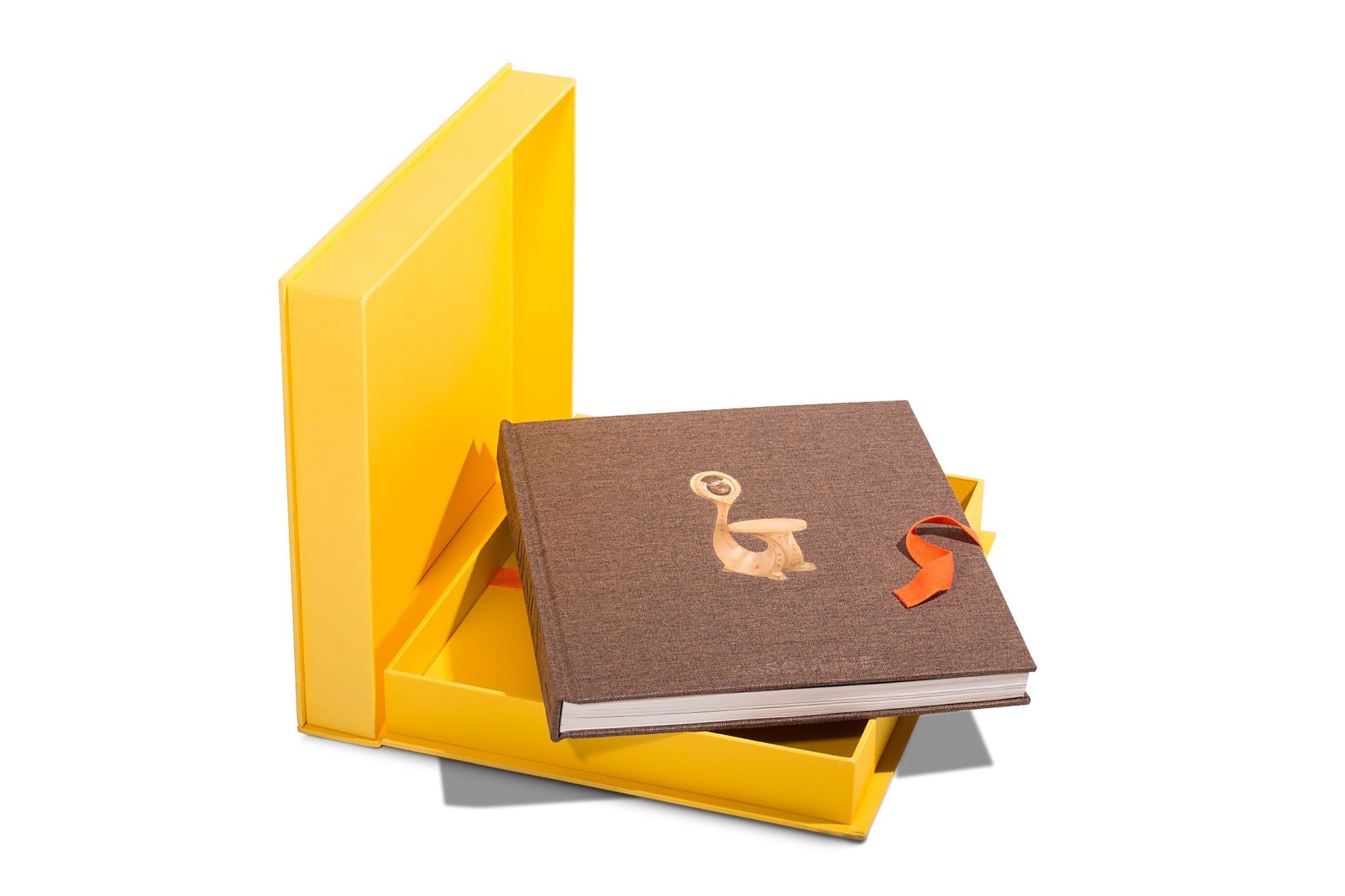 Assouline's "The Impossible Collection of Design" rests partially open in a yellow box. The brown hardcover features a circular cutout reminiscent of 20th-century design, with an orange ribbon extending from it.