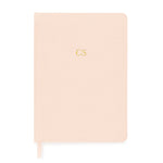 The Sugar Paper Tailored Pale Pink Journal is a closed hardcover with gold "CS" initials, a matching grosgrain ribbon bookmark, and lined pages for organized notes.