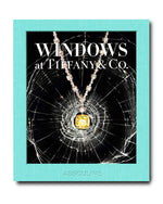The book cover, "Windows at Tiffany and Co." by Assouline, features a backdrop of shattered glass and highlights a yellow gemstone necklace, evoking Gene Moore's iconic Tiffany & Co. window displays.