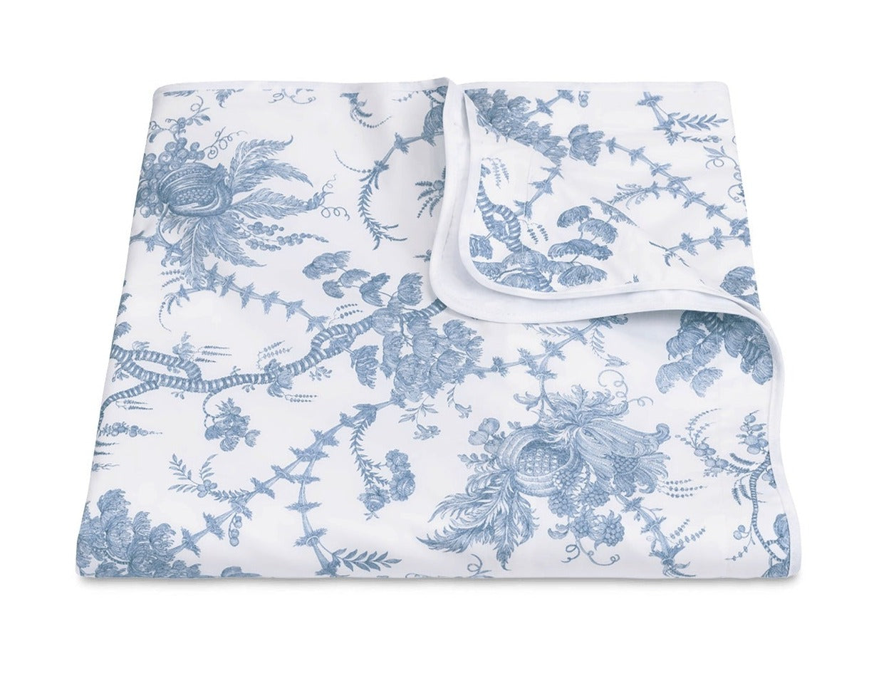 A folded white blanket from the San Cristobal Bedding Collection, Sky by Matouk, featuring a blue floral and botanical print on Luca Egyptian cotton percale, OEKO-TEX® Standard 100 certified, with one corner turned to show the reverse.