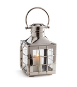 The Napa Home & Garden Nantucket Nickel Outdoor Lantern, Small, features glass sides, a handle, and a lit white candle inside, offering an elegant metallic accent to elevate your outdoor space.