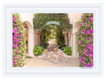 The Gray Malin Via Parigi Worth Avenue, Palm Beach photograph in a white frame (15.5” x 21”) features an archway adorned with pink flowers and vines, echoing Worth Avenue's elegance, with two orange bikes and white tires akin to a timeless capture awaiting its moment.