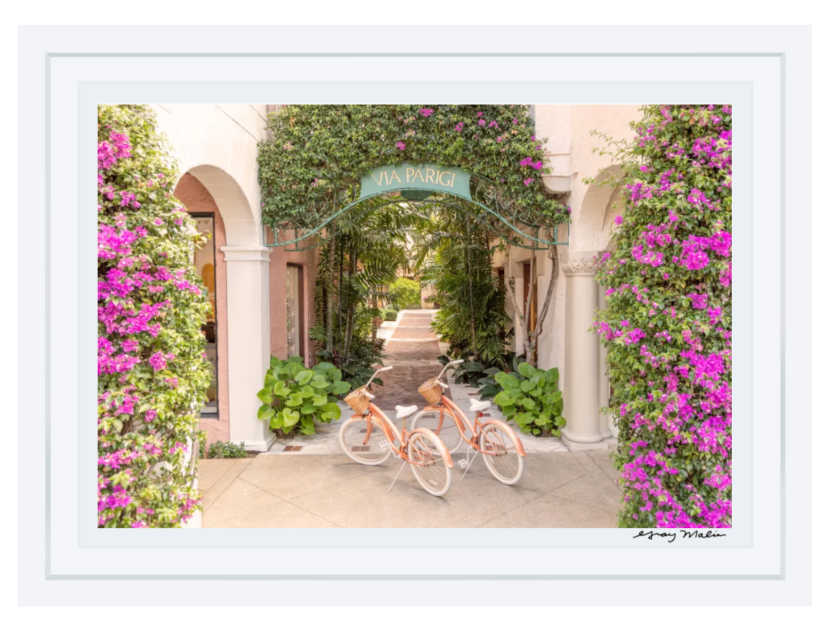 The Gray Malin Via Parigi Worth Avenue, Palm Beach photograph in a white frame (15.5” x 21”) features an archway adorned with pink flowers and vines, echoing Worth Avenue's elegance, with two orange bikes and white tires akin to a timeless capture awaiting its moment.