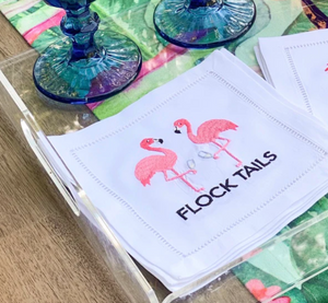 The August Morgan Cocktail Napkins, featuring embroidered pink flamingos and "FLOCK TAILS," are elegantly displayed on a wooden surface with vibrant glassware, making these cotton napkins an ideal unique gift.