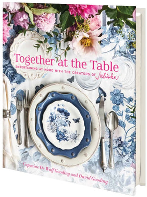 The book cover of "Juliska Together at the Table: Entertaining At Home With the Creators of Juliska" showcases an elegant dining scene with a floral plate setting enhanced by pink flowers on a signature blue Juliska-patterned tablecloth. This work by Capucine De Wulf Gooding and David Gooding invites you to celebrate life's moments.
