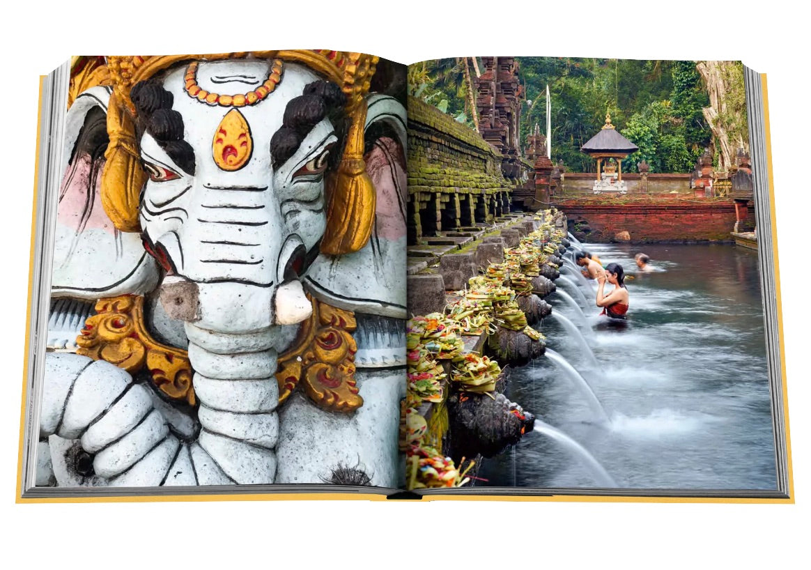 An open book displays two images: a close-up of an intricately decorated elephant statue and people bathing in a series of water spouts at a temple on the tropical paradise of Bali. The book is titled **Bali Mystique** by **Assouline**.