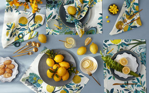 The table setting includes the Matouk Citrus Garden Pool Napkin Set with floral themes, Josef Frank-inspired plates, cutlery, and glasses of lemon water. Lemons and yellow candies are scattered across the table, creating a vibrant and picturesque arrangement.