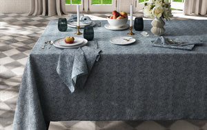 A dining table set with Matouk’s Rubia Navy Napkin Set, plates, apples, glassware, candles, and a vase of white flowers in a room with checkered floor tiles and large windows. OEKO-TEX® Standard 100 certified.