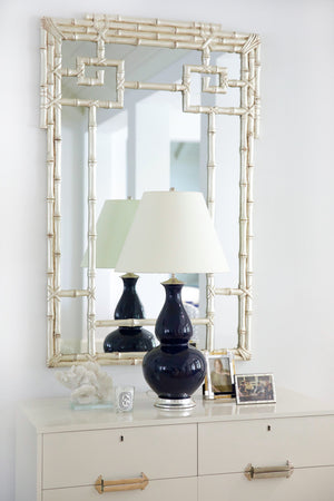 A white dresser with gold handles displays a navy blue lamp, framed photos, and coral decor, set against the Carvers' Guild Bamboo Mirror in Silver featuring an elegant geometric motif.