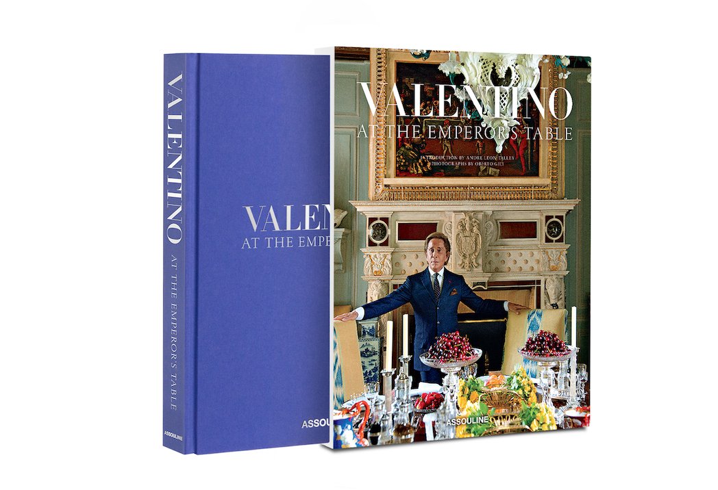 The Assouline book "Valentino: At the Emperor's Table" has a vibrant cover with haute couture flair, displaying a lavish dining setup and a man in a suit. Beside it, a plain blue hardcover contrasts yet complements the elegance of Valentino Garavani's world.