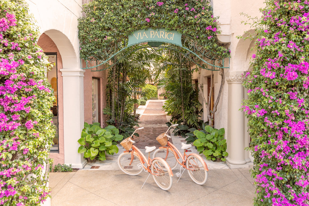 Two bicycles with baskets sit under a "Via Parigi" archway amid greenery and purple flowers in Palm Beach, ideal for Gray Malin's lens. Buy the Gray Malin Via Parigi, Worth Avenue, Palm Beach, 15.5” X 21” with White Frame to bring this charm home.