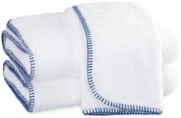 A stack of white fluffy Matouk Whipstitch Bath Collection - Periwinkle towels with blue whipstitch embroidered edges.