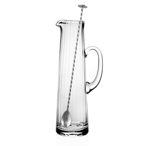 The Corinne Tall Cocktail Jug & Spoon by William Yeoward Crystal is a handmade, clear glass jug with a handle, paired with a long, twisted metal stirring spoon.