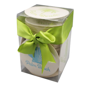 A clear box of round Palm Beach Coasters by Haute Papier, elegantly packaged with a wide green ribbon and bow—makes a perfect hostess gift.