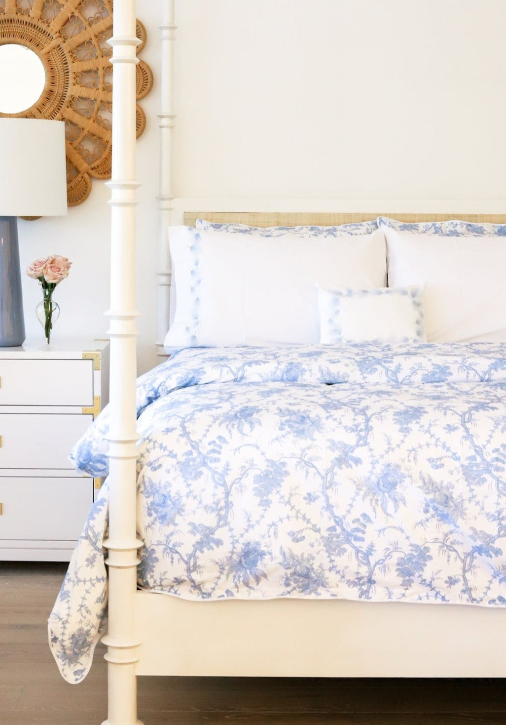 A neatly made bed with the Matouk San Cristobal Bedding Collection in Sky, white pillows, and a small decorative cushion; a white nightstand with a lamp, flowers, and a mirror is beside the bed.