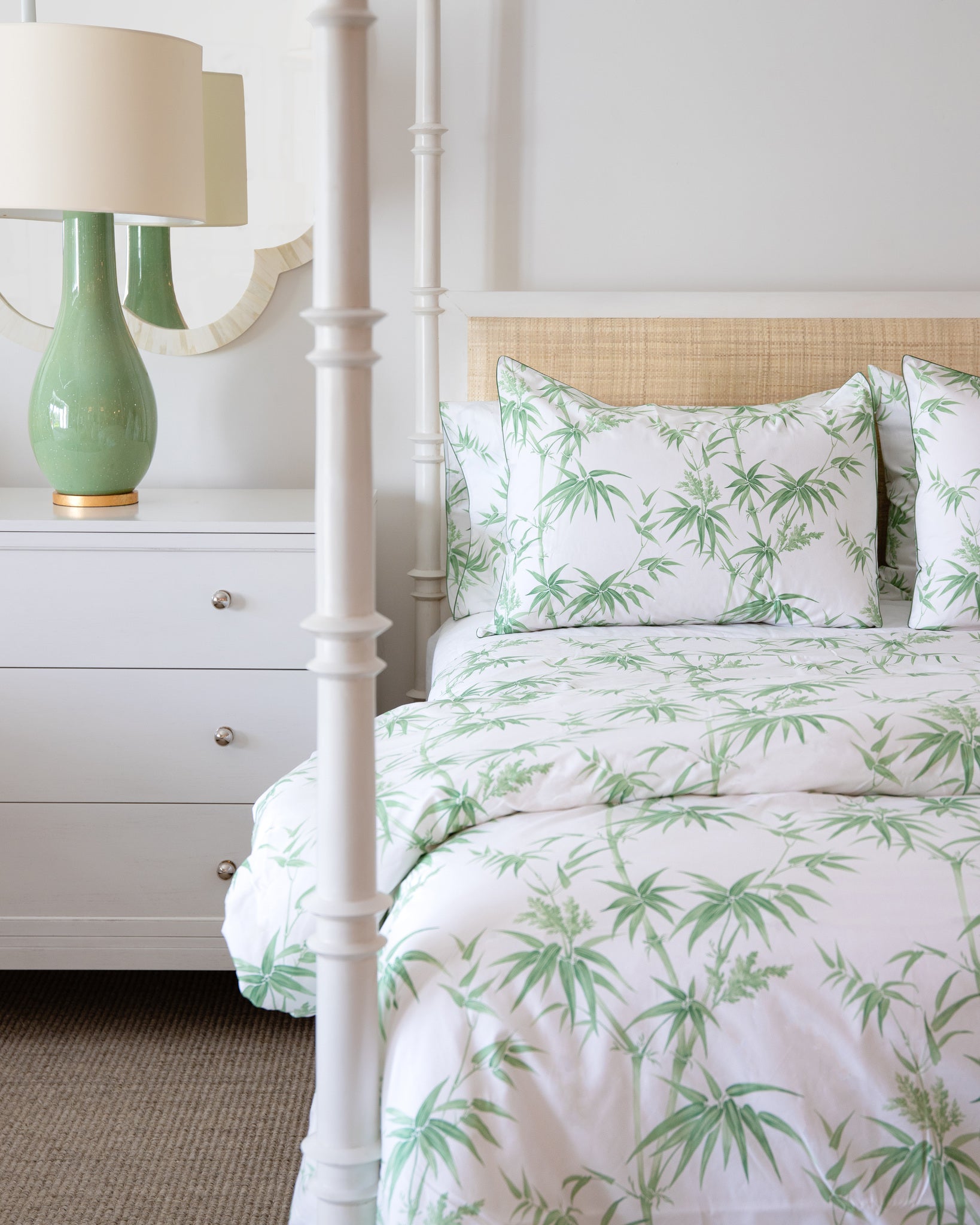 A neatly made bed features the Matouk Dominique Bedding Collection in a green palm pattern, crafted from Egyptian cotton, next to a white dresser with a green lamp and round mirror in this bright, airy bedroom.
