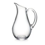 The Woodstock Large Glass Pitcher by Simon Pearce features a wide base and curved handle—its clear glass design makes it an elegant vessel, shown empty against a plain white background.