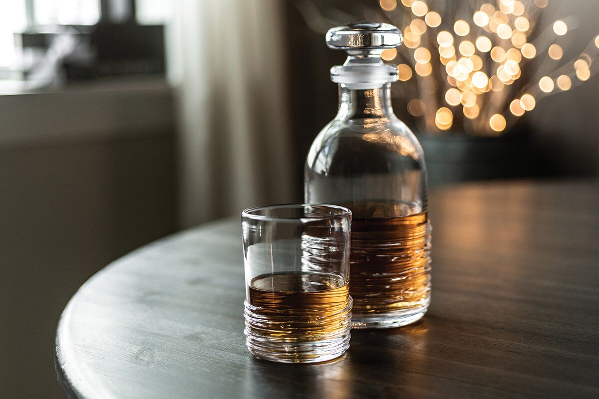 The Simon Pearce Echo Lake Decanter and glass, both filled with amber liquor, sit on a wooden table against blurred lights, highlighting the craftsmanship of master glassblowers.
