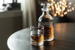 The Simon Pearce Echo Lake Decanter and glass, both filled with amber liquor, sit on a wooden table against blurred lights, highlighting the craftsmanship of master glassblowers.