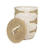 The Jelsa Round Hamper by Pigeon & Poodle is crafted from natural and white raffia in a geometric pattern with a removable lid, shown here against a white background.