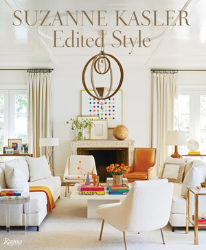 The signature interior design of "Suzanne Kasler: Edited Style" by Rizzoli showcases an elegant living room with modern white and brown furniture, vibrant books, and a decorative chandelier.