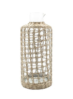 The Be Home Seagrass Cage Carafe features a clear cylindrical design with seagrass wrapping, a small spout, and an open top. Handwash is recommended to preserve its beauty.