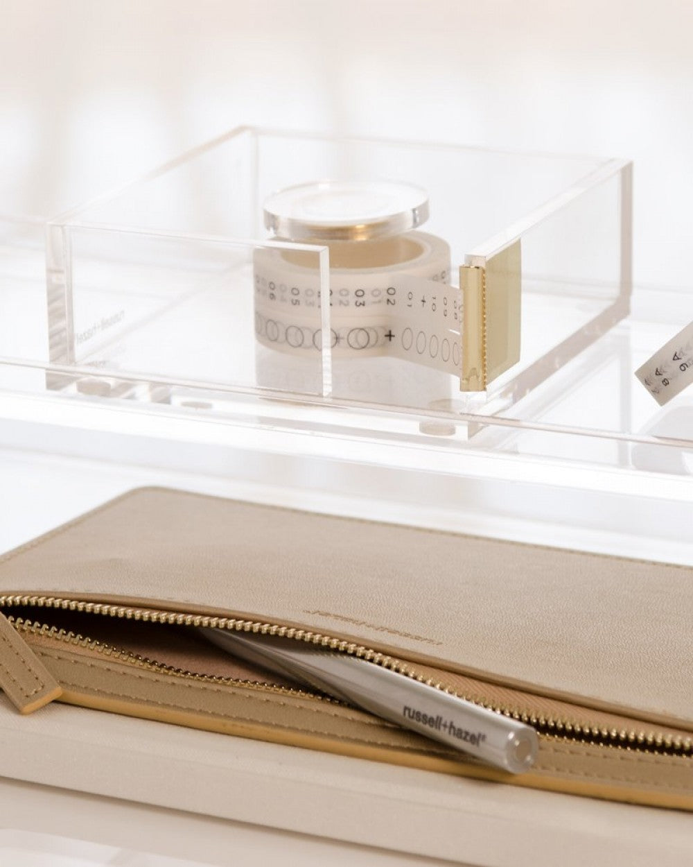 The Russell & Hazel Acrylic Tape Bloc, a clear acrylic and gold tape dispenser, rests atop a beige zippered case with a branded pen—a sleek addition to any desktop organizer.