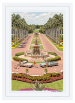 Gray Malin’s The Main Drive, The Breakers Palm Beach (24" x 36", White Frame) captures a vibrant scene with statues, vintage cars, and brightly dressed people along a palm-lined driveway at The Breakers Palm Beach.
