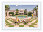 Two people relax on lounge chairs beside a pool at POOLSIDE AT LA FOLLIA, surrounded by greenery and sculptures. The Palm Beach architecture adds elegance to this Gray Malin Poolside at La Follia, Palm Beach scene in a White Frame, 29.5" x 41.5".