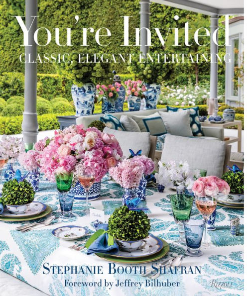 A beautifully arranged outdoor dining table adorned with pink flowers, a blue and white patterned tablecloth, and vibrant glassware awaits guests amidst lush greenery. This idyllic setting for entertaining at home is showcased on the cover of "You’re Invited: Classic, Elegant Entertaining" from Common Ground.